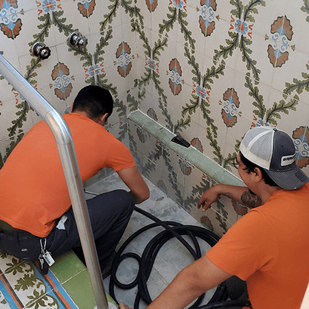 Two technicians working together to inspect or repair a drain in a tiled shower area.
