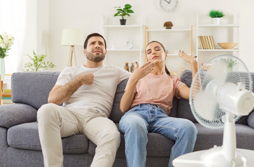 A young couple sitting in front of a fan, sweating.